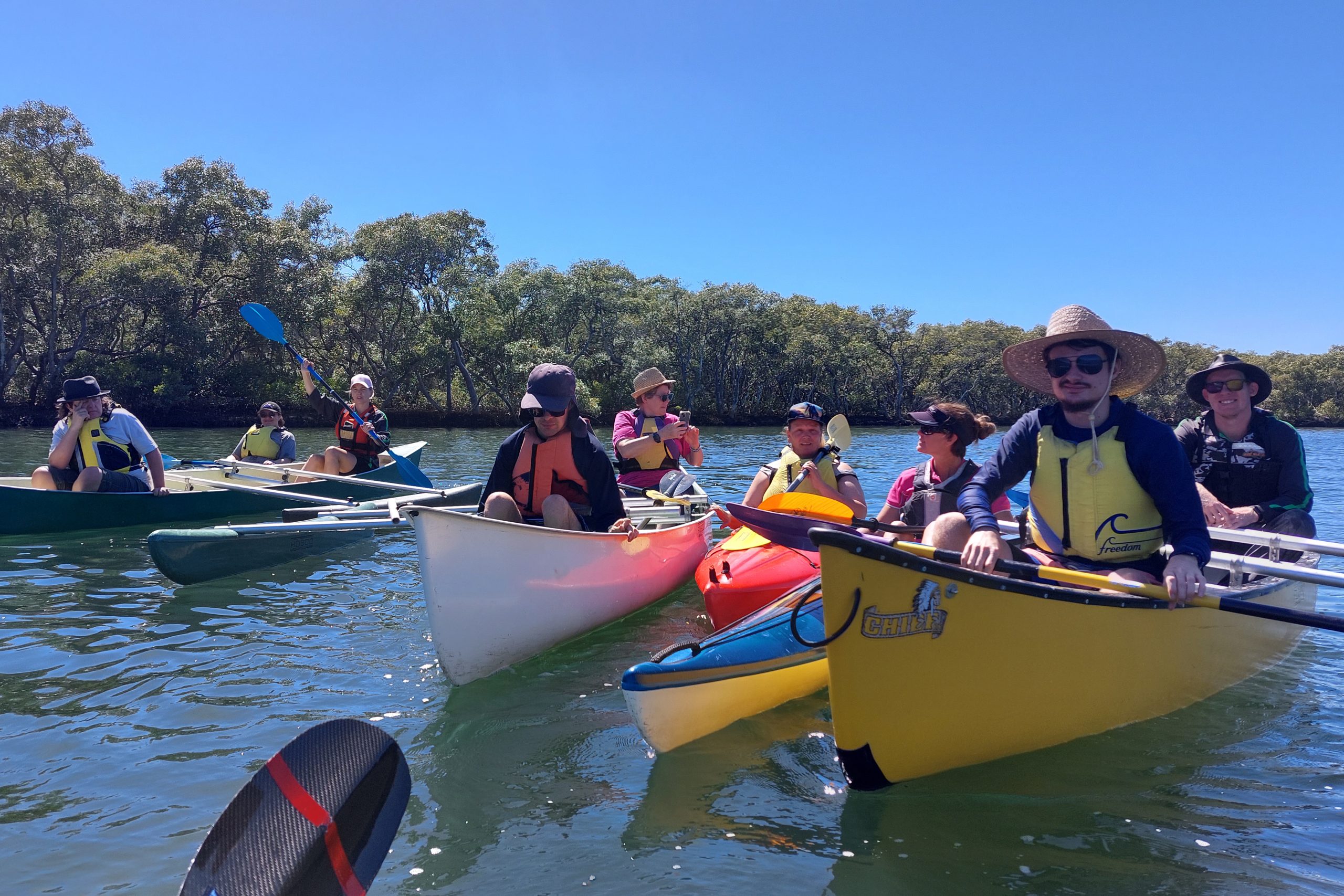 A group of nine people in hats and life jackets sitting in five different canoes and kayaks on the water under a clear blue sky.