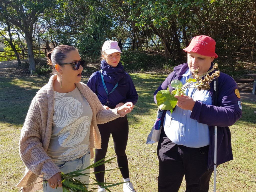 A man with a white cane examining some large leaves and open seedpods on a stick, as a Quandamooka woman tells him about their history and uses, and another young woman watches on.