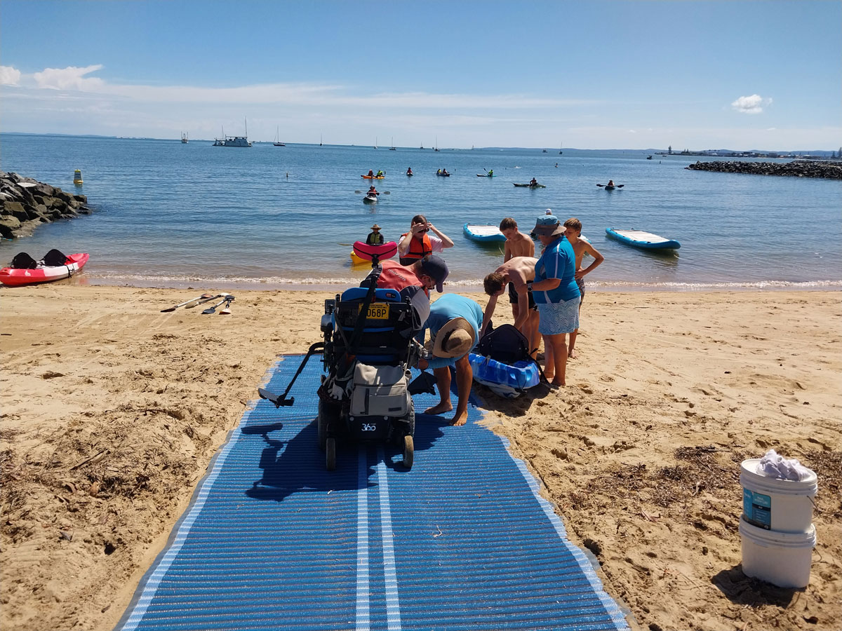Sunny day at a beach with a blue wheelchair ramp in use down to the water with multiple people assisting 