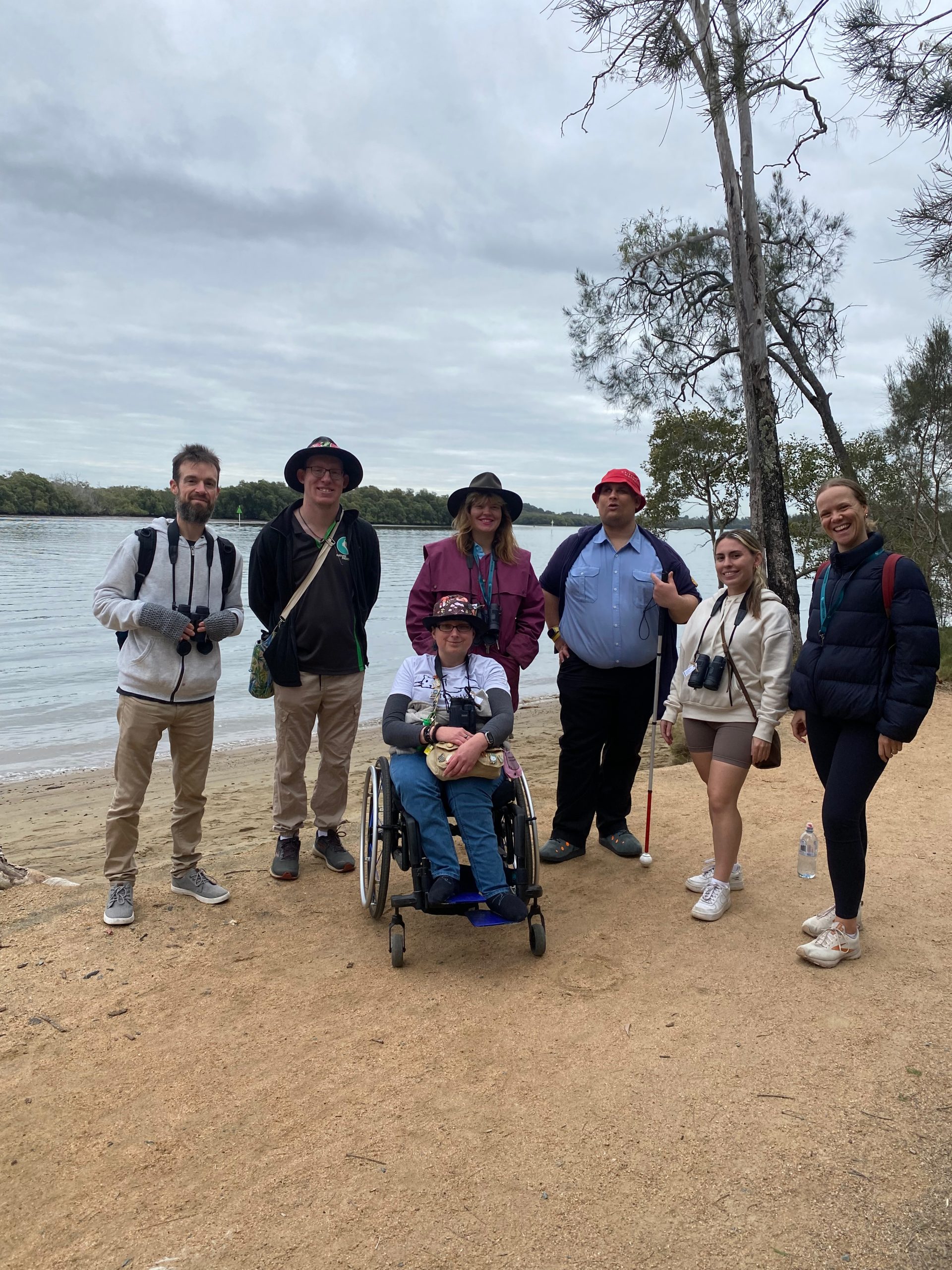 A group of six smiling people, including a woman in a wheelchair, standing on a sandy shoreline near a couple of gumtrees.